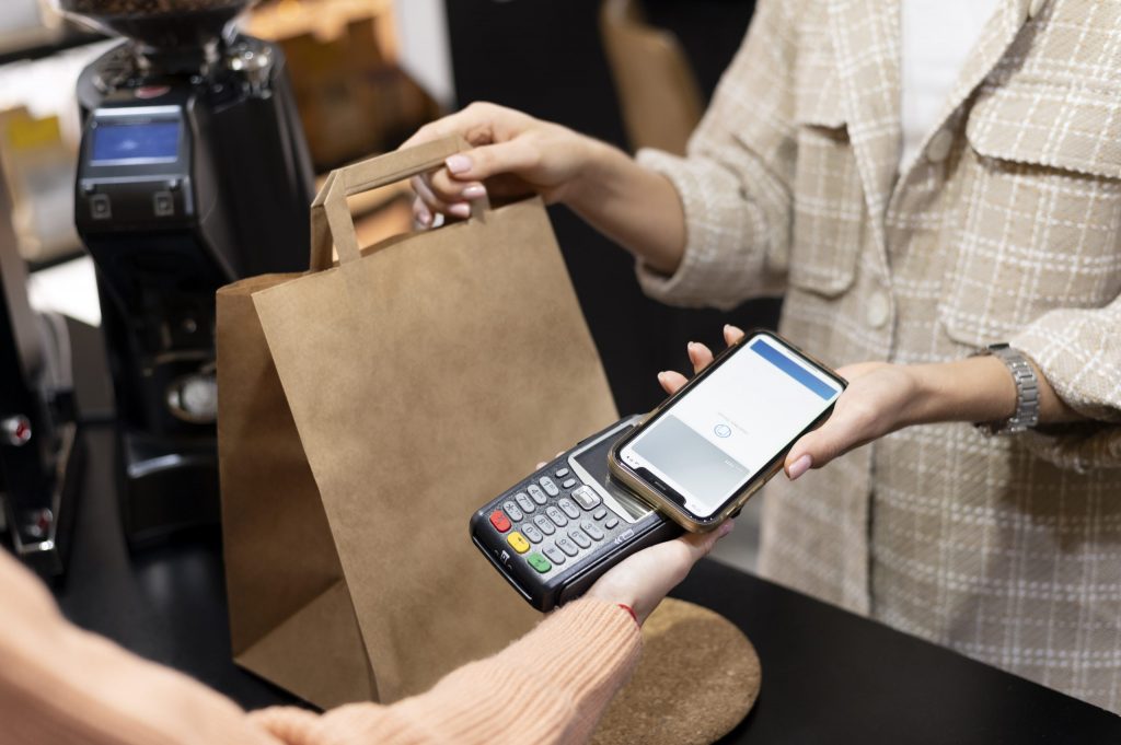 Eine Person bezahlt kontaktlos mit ihrem Smartphone an einem Schalter, während ihr eine andere Person eine braune Papiertüte reicht.